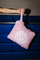 A pink quillow with a volleyball graphic and a top handle, held by a person's arm in front of blue bleachers