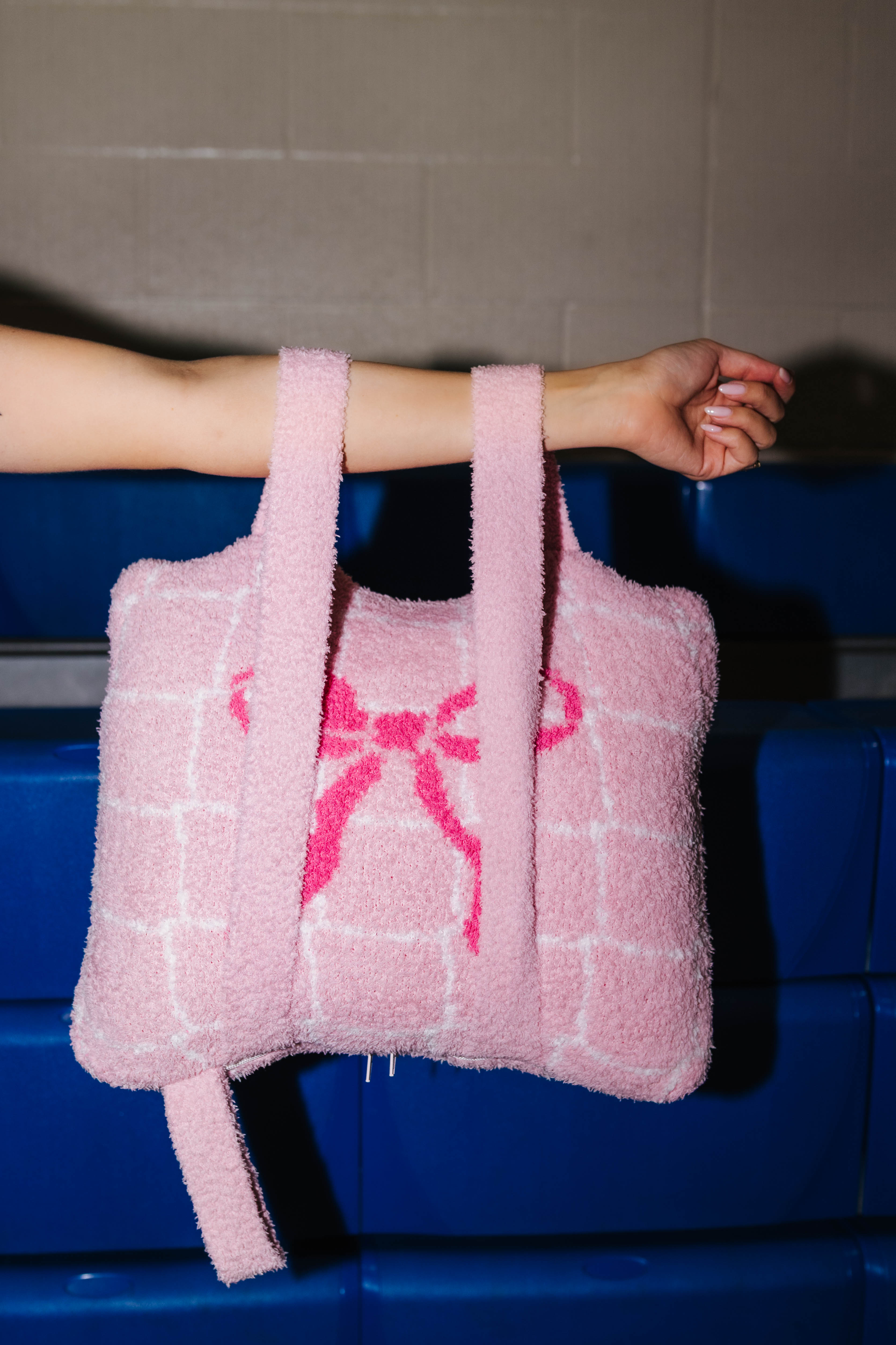 A pink quillow with a grid pattern and a pink bow design, held by a person's arm against a blue bench and gray wall background.