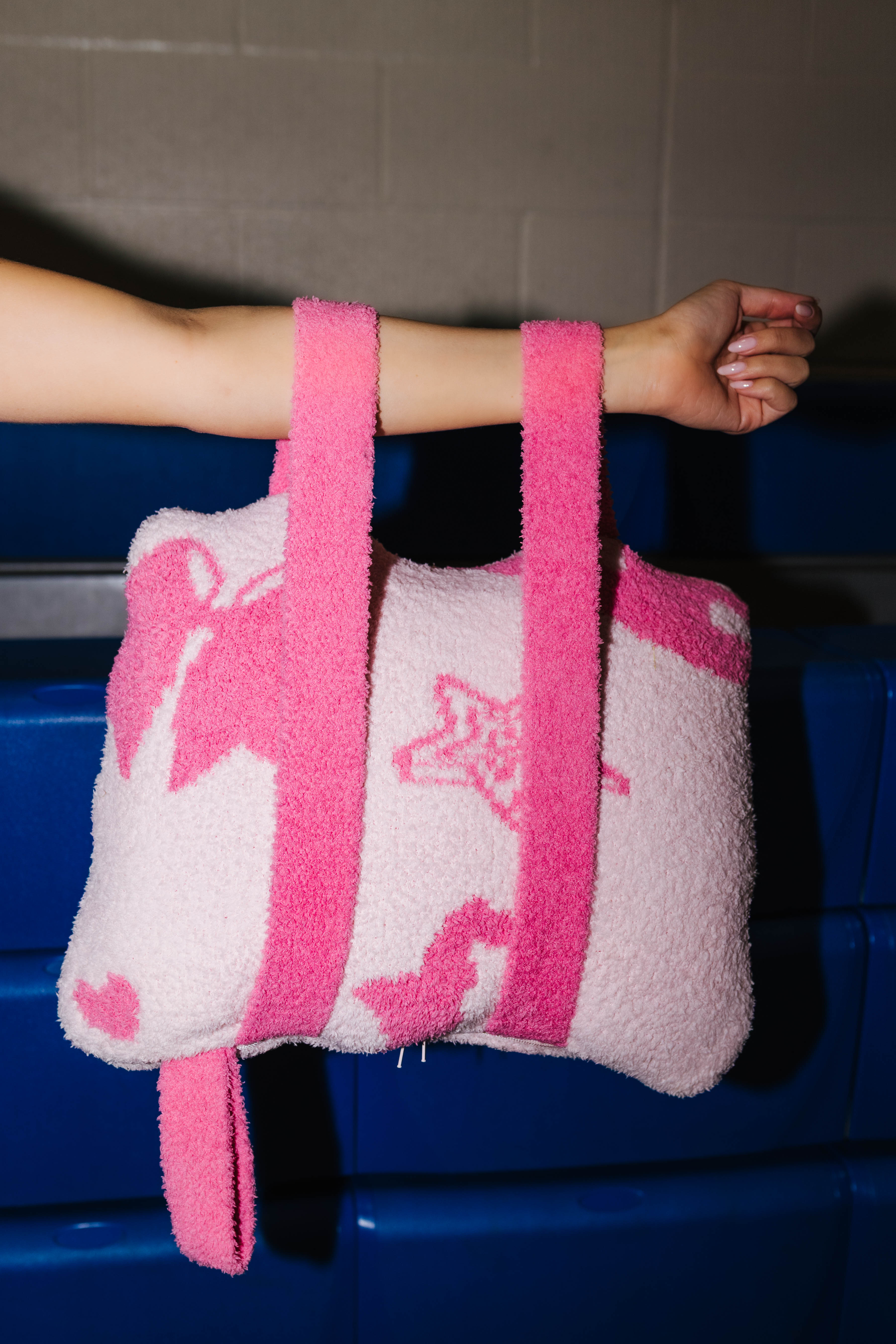 A fuzzy quillow with pink straps, held by a person's arm against a dark blue bench and gray wall background.