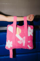 Holding the quillow by the backpack straps showing the white floral and bow patterns, held by a person's arm against a dark blue and gray background.