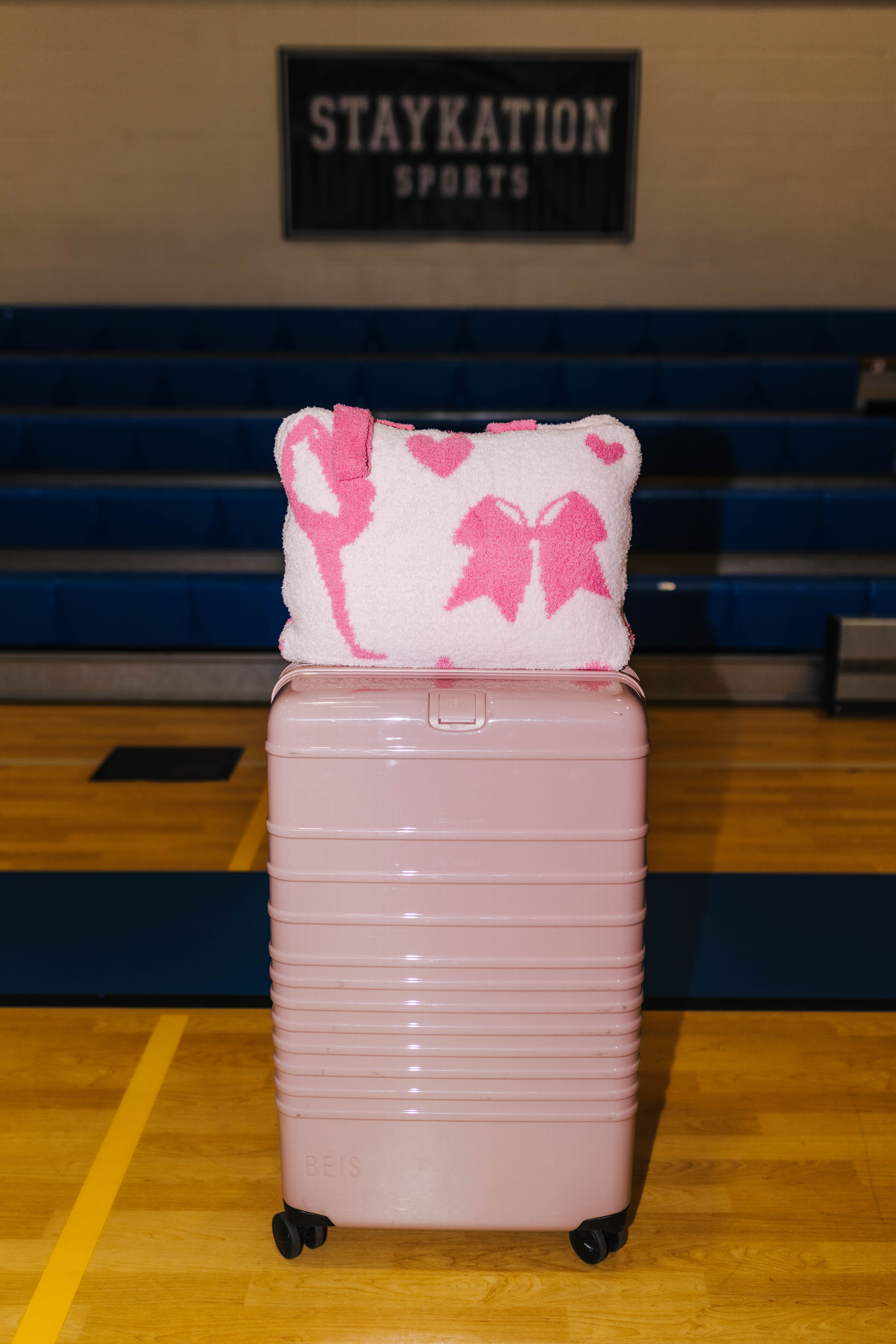 A light pink hard-shell rolling suitcase stands on a wooden gym floor in front of blue bleachers, with a white and pink patterned cheer quillow on top. 