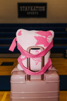 A pink and white patterned quillow on a light pink hard-shell suitcase, set in front of blue stadium seating with a 'STAYKATION SPORTS' sign in the background.