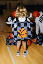 A child stands on a basketball court wearing a black and gray checkered backpack quillow with a large orange basketball graphic on the back.