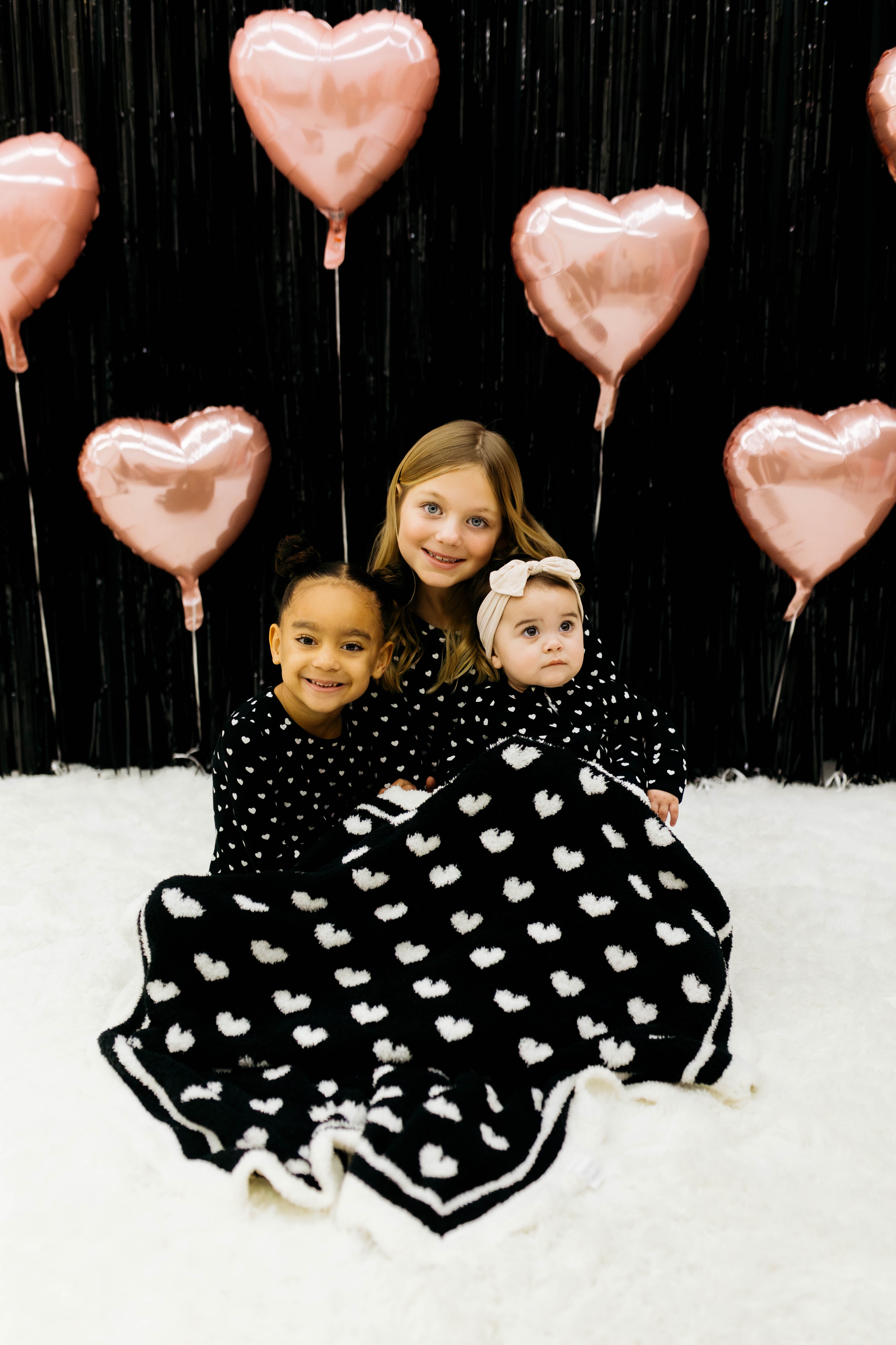 Three children in matching black and white pjs sitting on a white surface with pink heart balloons in the background. With the black blanket draped on them.