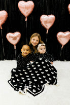 Three children in matching black and white pjs sitting on a white surface with pink heart balloons in the background. With the black blanket draped on them.