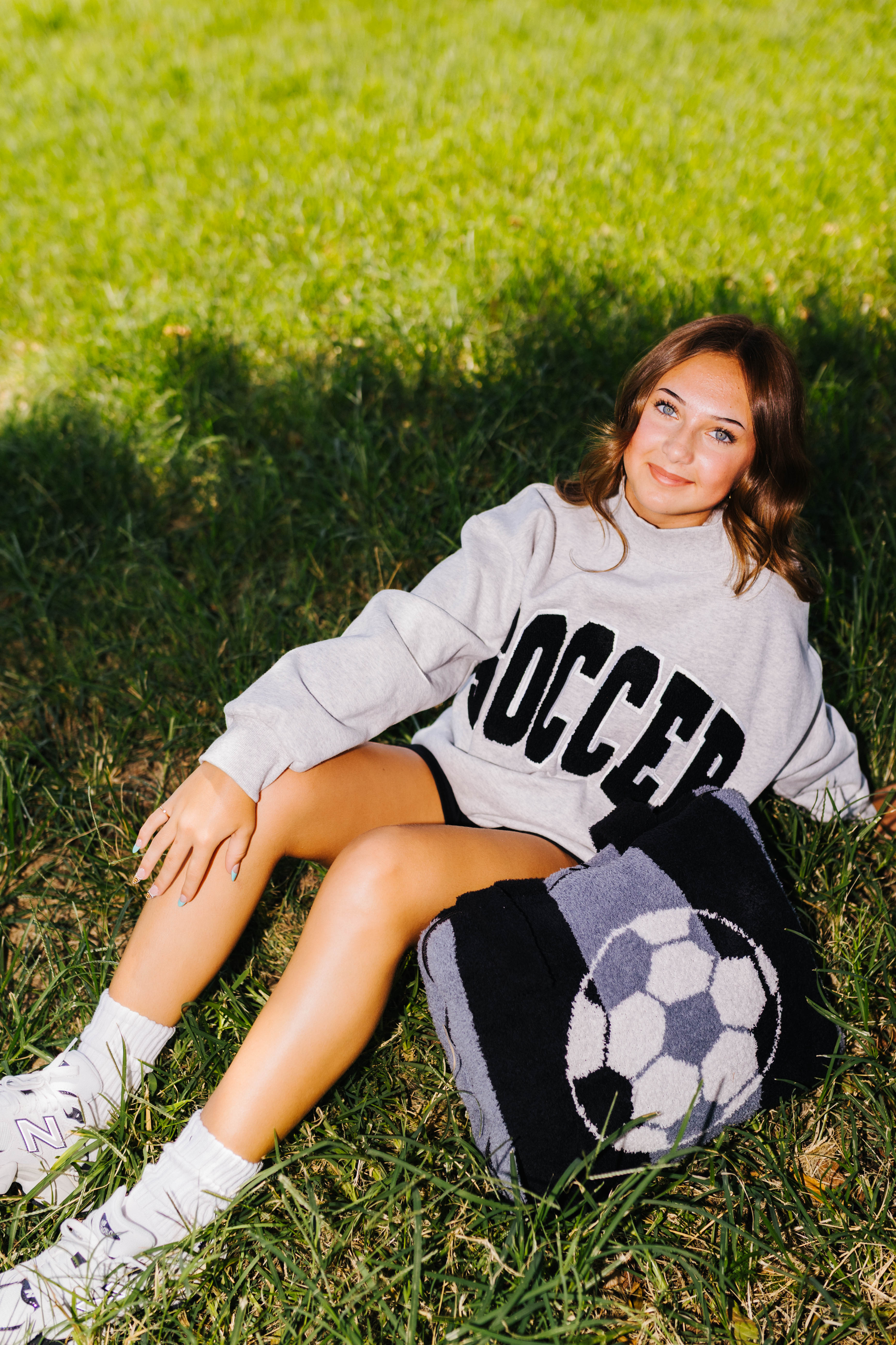 A young woman sits on a grassy lawn wearing a light gray sweatshirt with 'SOCCER' printed in black, black shorts, and white sneakers. A black and white soccer-themed knit quillow rests beside her.