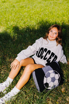 A young woman sits on a grassy lawn wearing a light gray sweatshirt with 'SOCCER' printed in black, black shorts, and white sneakers. A black and white soccer-themed knit quillow rests beside her.