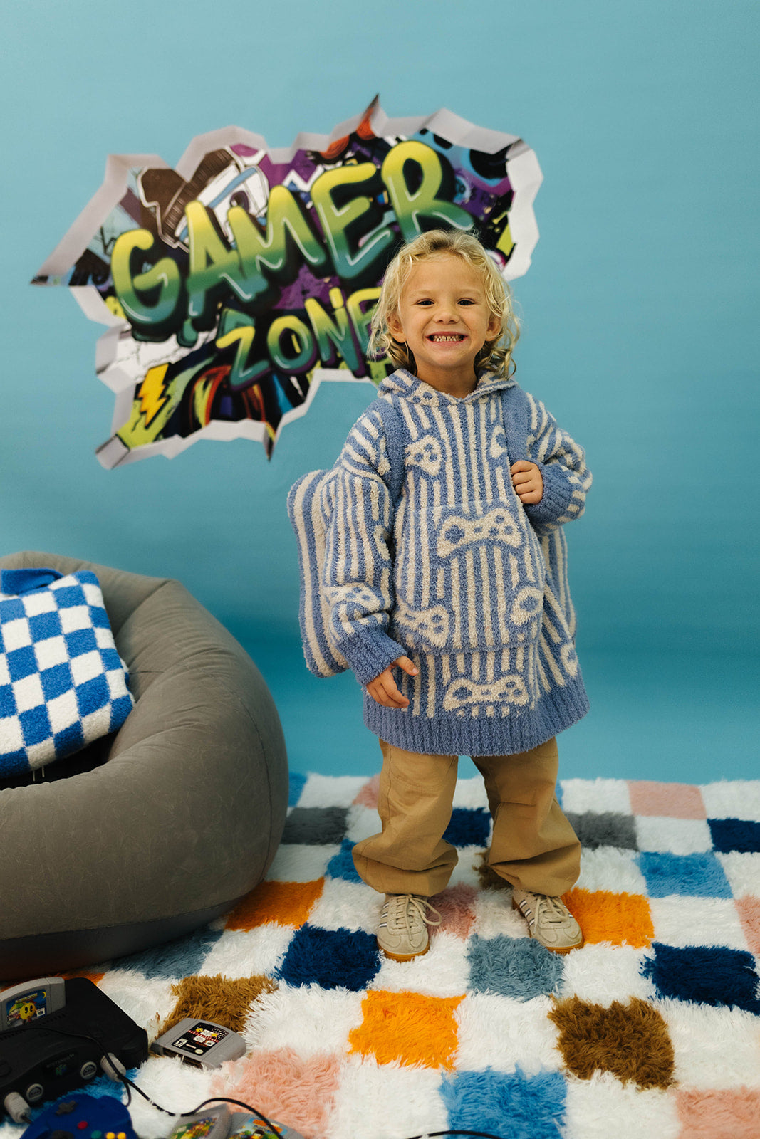 Child in a patterned sweater standing in front of a 'Gamer Zone' sign with a colorful rug and bean bag chair.