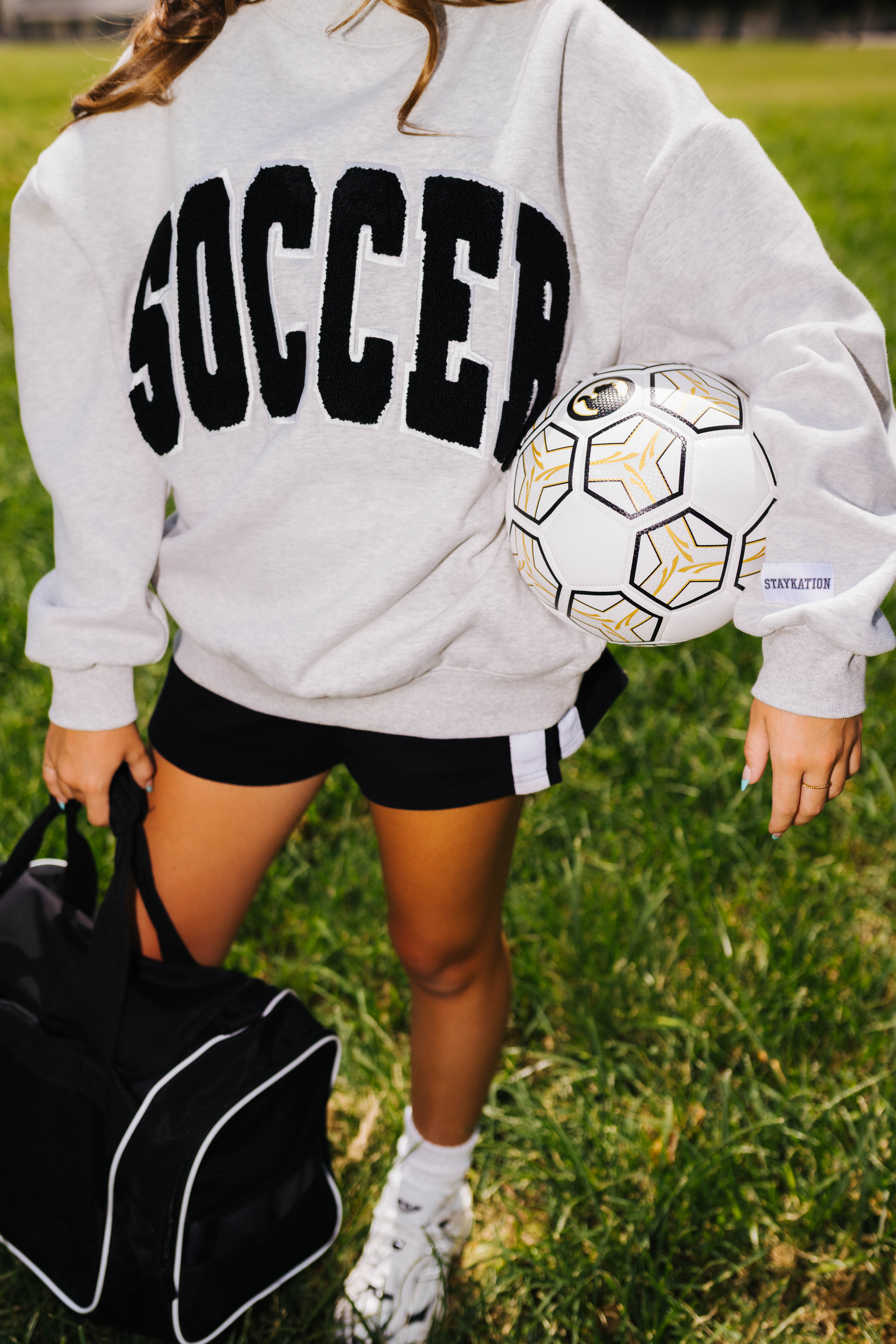 A person outdoors on grass wearing a light gray sweatshirt with large black 'SOCCER' text across the chest, black athletic shorts, white socks, and holding a soccer ball and a black duffel bag.