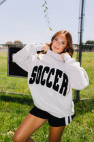 A woman posing outdoors in a gray crewneck sweatshirt with 'SOCCER' printed in black across the chest, paired with black athletic shorts, in front of a chain-link fence and grassy field.