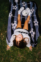 A girl lies on her back on green grass, wearing a gray sweatshirt, black shorts, and white socks, with a soccer ball on her feet. She is lying on a black and gray striped blanket with soccer ball patterns.