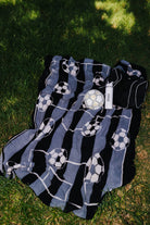 A black and blue striped blanket with white soccer ball patterns, laid out on green grass next to a white soccer ball.