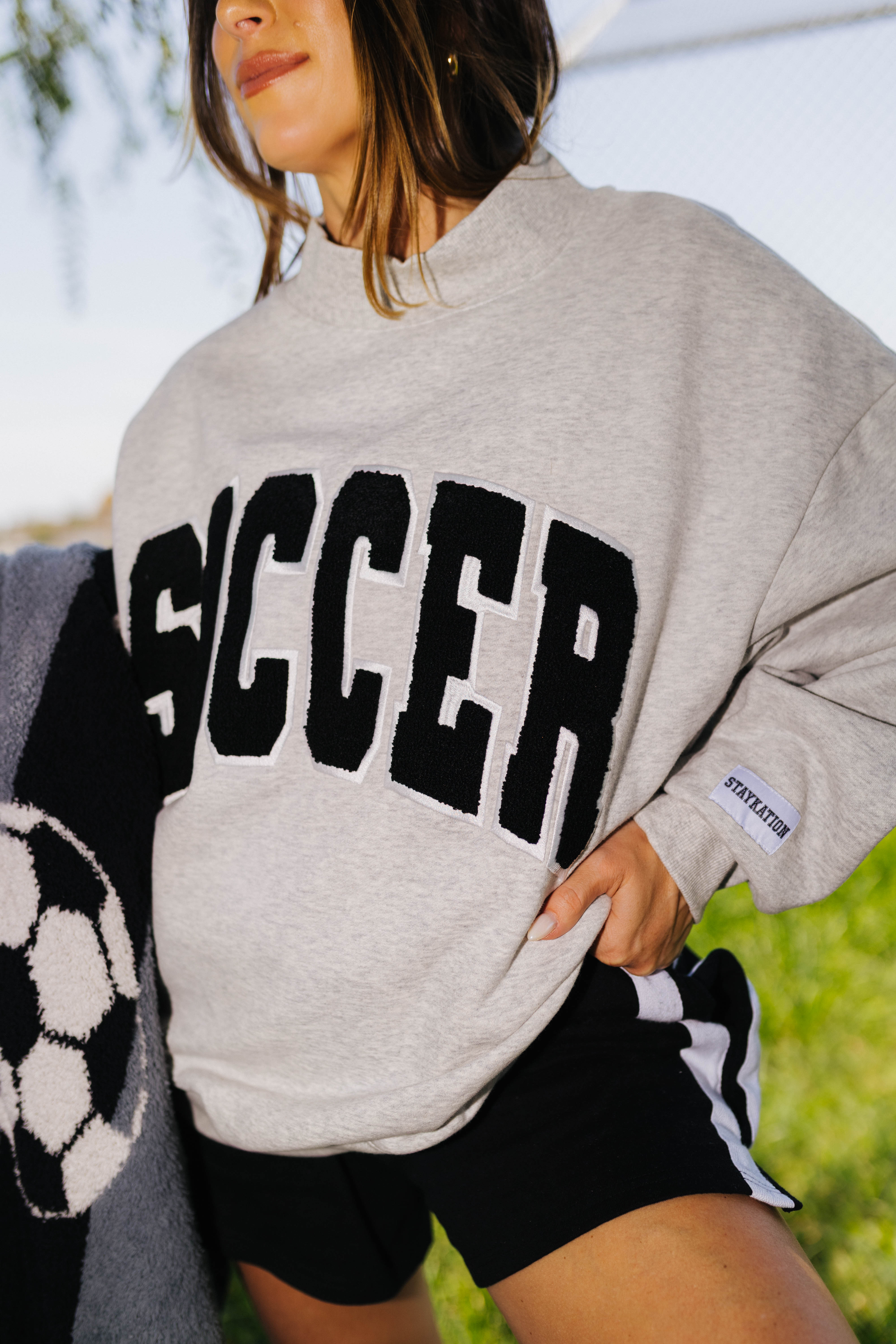 A woman wearing an oversized light gray knit sweatshirt with the word 'SOCCER' in large black varsity-style letters across the front, paired with black athletic shorts with white side stripes. The photo is taken outdoors with a blurred green and blue background.