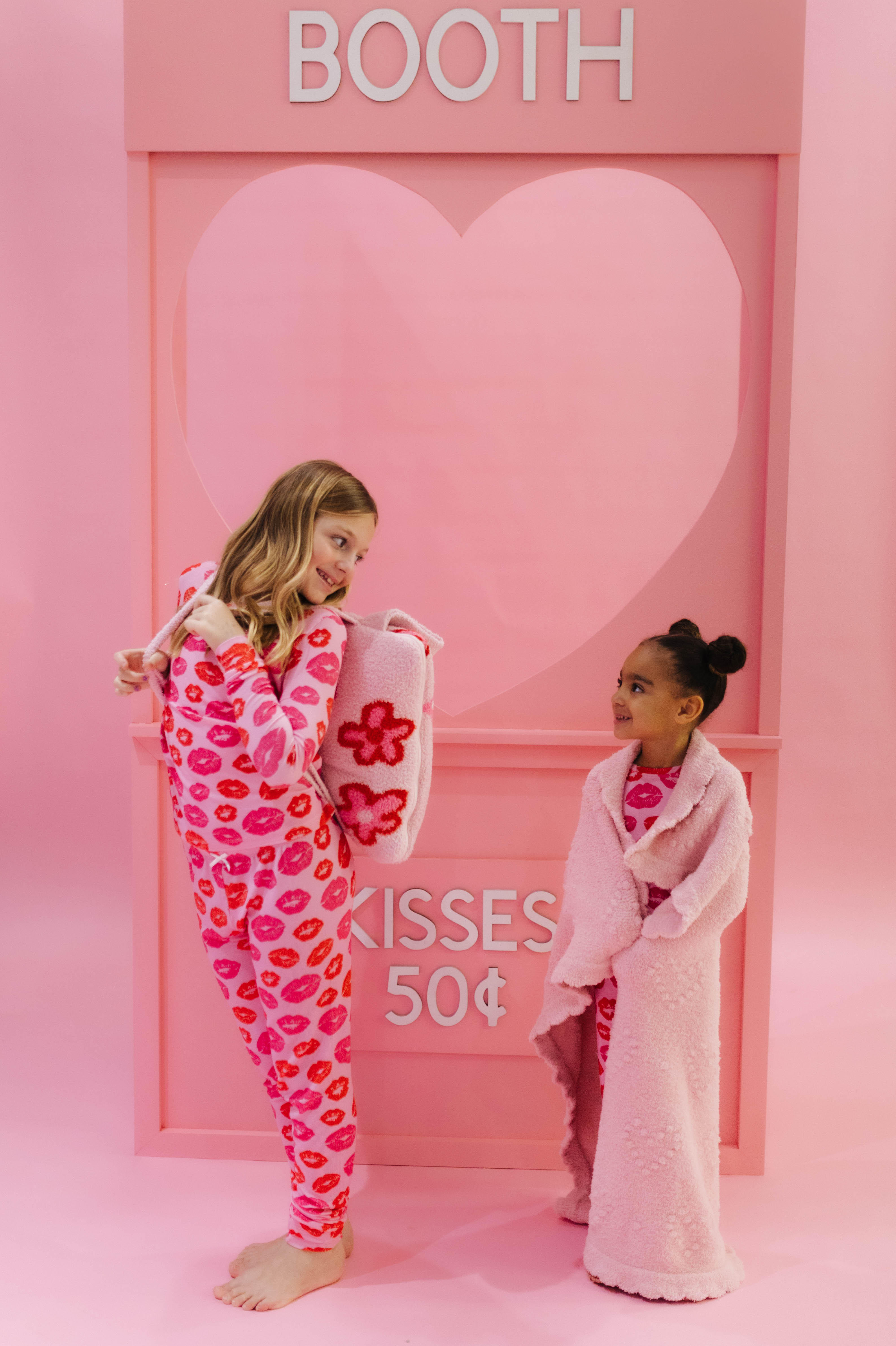 Two children in pink-themed outfits standing in front of a heart-shaped pink backdrop with 'BOOTH' and 'Kisses 50c' text.