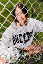 A woman sitting on grass behind a chain-link fence, wearing a light gray sweatshirt with 'SOCCER' printed in black across the front and holding a soccer ball.