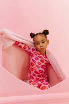 Child wearing a pink outfit with red patterns, sitting on a pink surface. Holding up the pink heart blanket