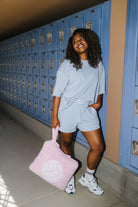 A woman stands in a hallway with blue lockers, wearing a light blue oversized t-shirt and matching shorts, holding a pink volleyball-themed quillow.