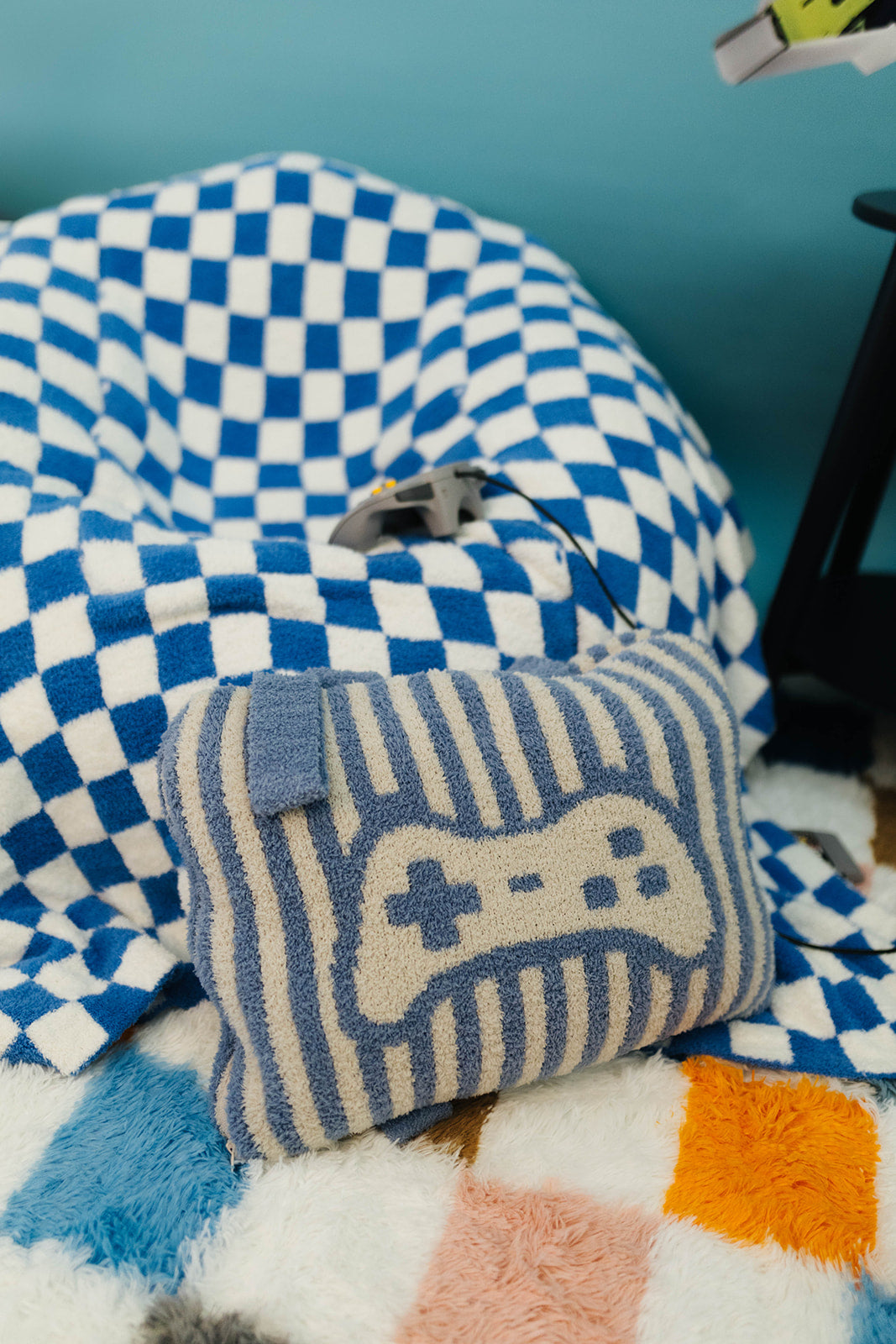 Blue and white checkered bean bag chair with a striped pillow featuring a game controller design on a colorful rug.