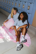 Two women sitting on a pink blanket in front of blue school lockers, wearing matching light blue t-shirts and shorts.