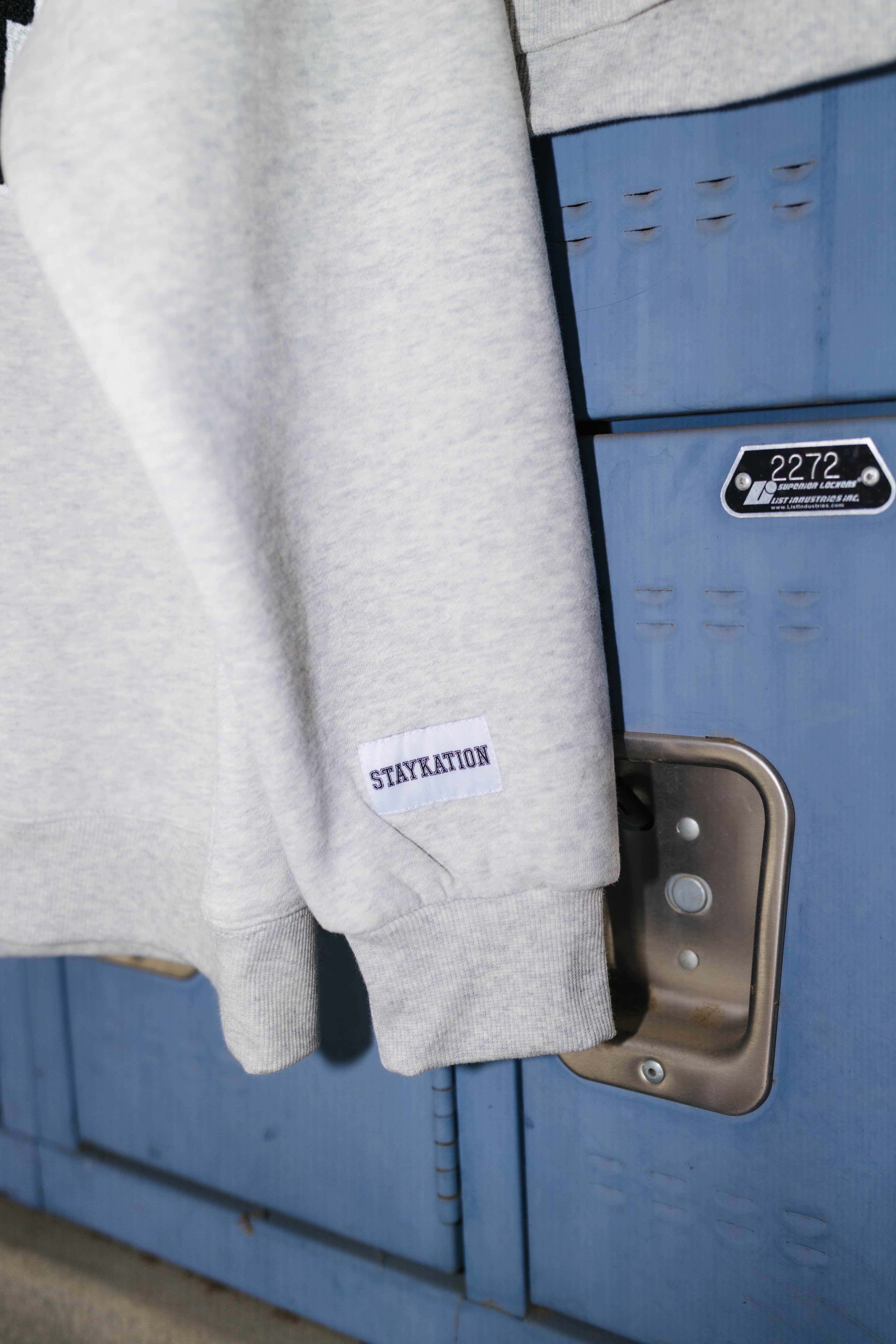 A light gray heather sweatshirt draped over blue lockers, featuring a small white rectangular patch on the sleeve with the text 'STAYKATION'.