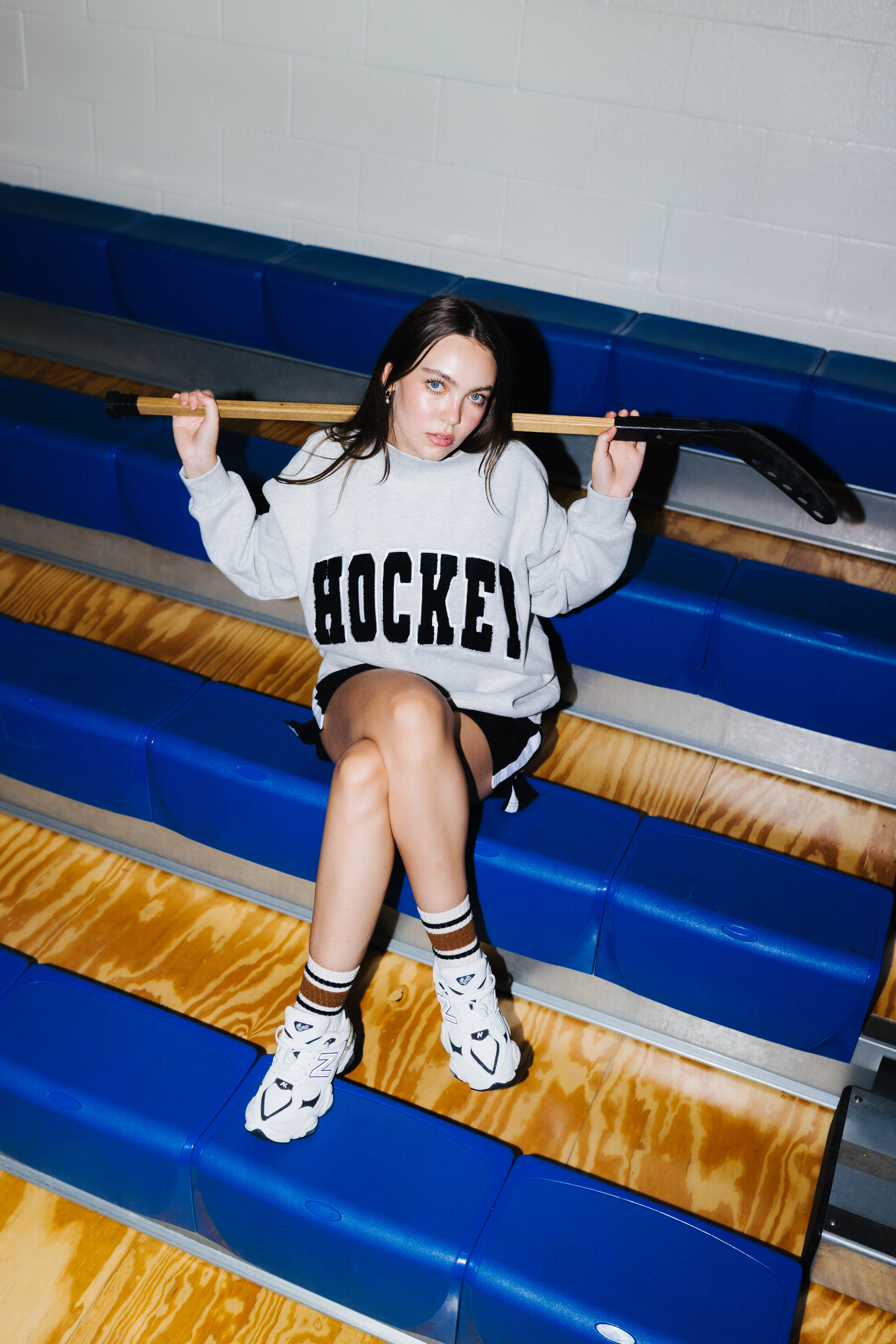 A woman sits on gym bleachers holding a hockey stick, wearing a light gray sweatshirt with 'HOCKEY' printed in black across the chest, black shorts, white socks, and white sneakers.