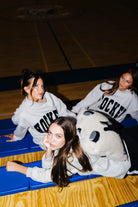 Three women in matching light gray crewneck sweatshirts with 'HOCKEY' printed in black across the chest, posed on blue gym bleachers in a basketball court.