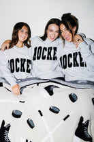 Three women sitting together on bleachers wearing matching light gray hoodies with 'HOCKEY' printed in black varsity-style letters across the front. A cream knit blanket with black hockey motifs is spread across the bed.