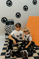 Two children sitting on a checkered floor with a wall featuring smiley face designs.