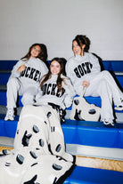 Three women sitting on blue gym bleachers wearing matching gray hockey sweatpants with black HOCKEY lettering with the blanket draped over them