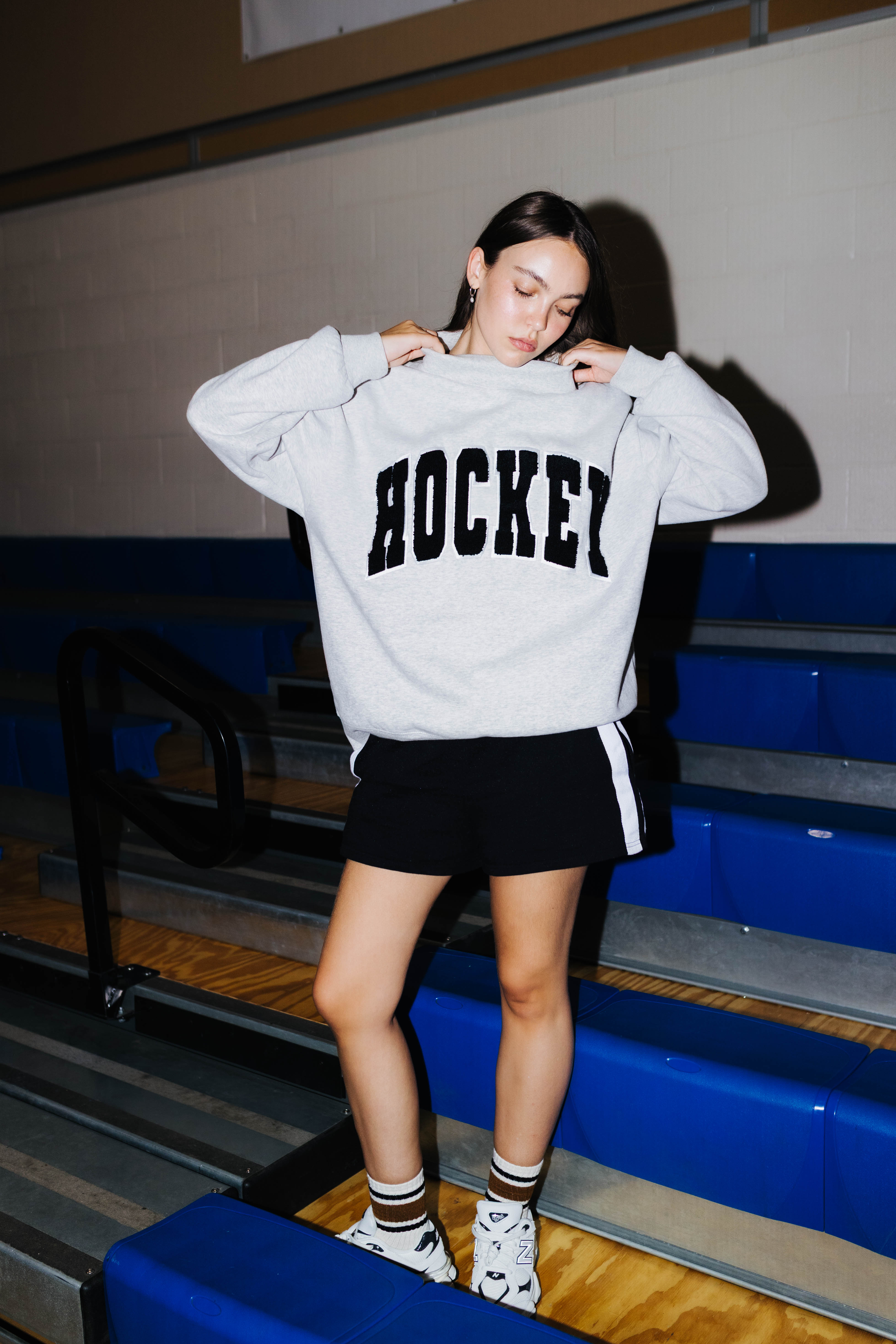 A woman wearing a light gray oversized sweatshirt with 'HOCKEY' printed in black across the chest, paired with a black shorts standing on blue bleachers in a gym.