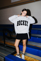 A woman wearing a light gray oversized sweatshirt with 'HOCKEY' printed in black across the chest, paired with a black shorts standing on blue bleachers in a gym.
