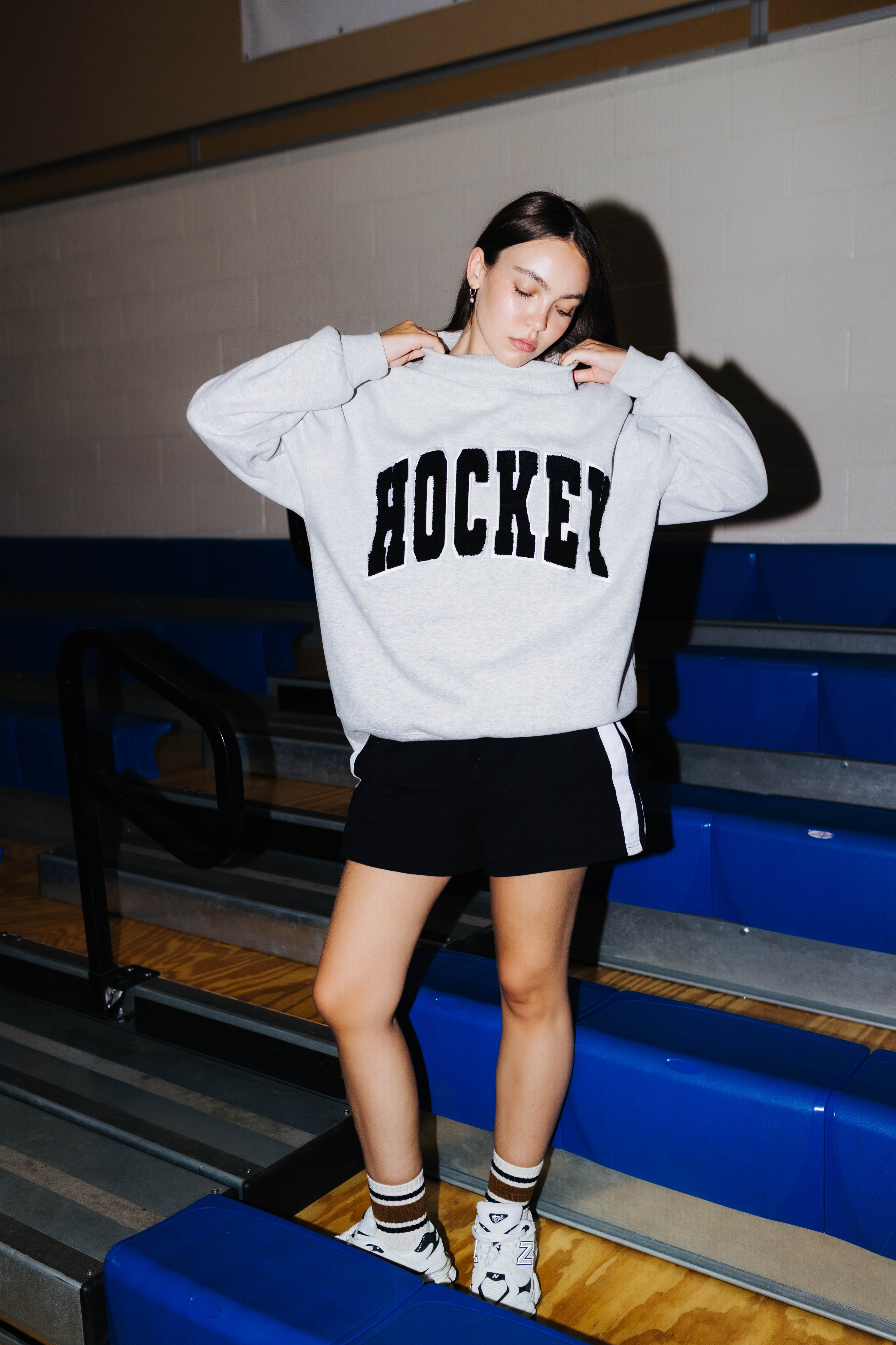 A woman wearing a light gray oversized sweatshirt with 'HOCKEY' printed in black across the chest, paired with a black shorts standing on blue bleachers in a gym.