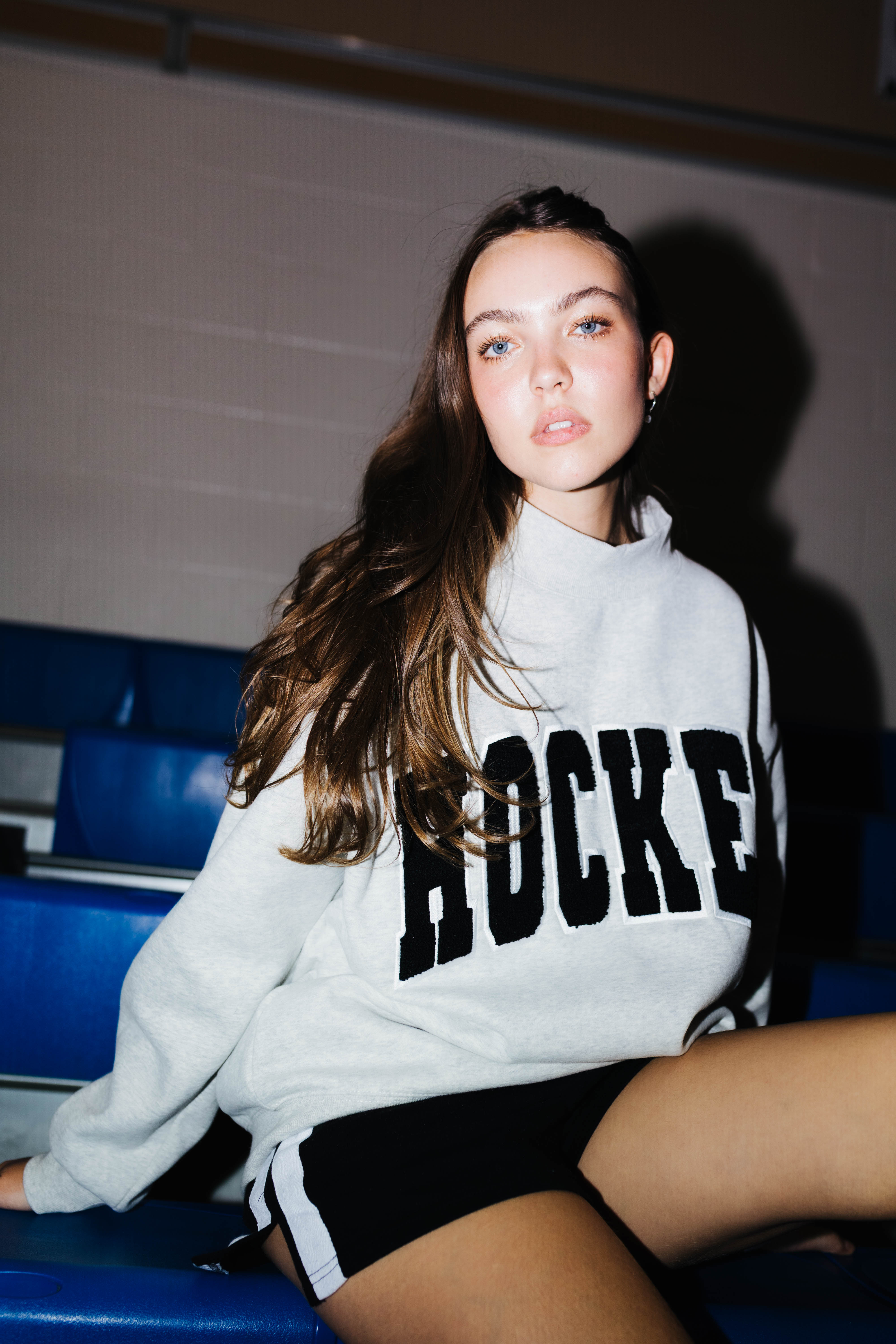 A woman sitting on blue stadium seating wearing a light gray sweatshirt with 'HOCKEY' printed in black across the chest, paired with black shorts.