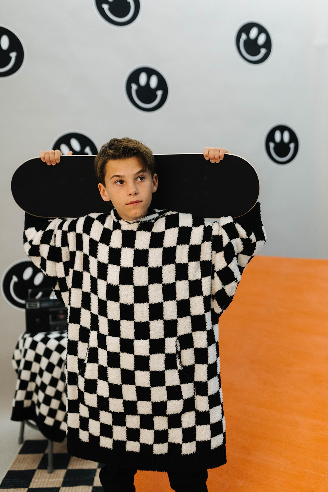 Person wearing a black and white checkered outfit holding a skateboard against a wall with smiley face designs.