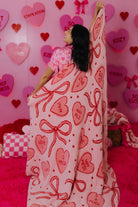 Person wrapped in a blanket with heart and bow patterns against a pink heart-patterned wall.