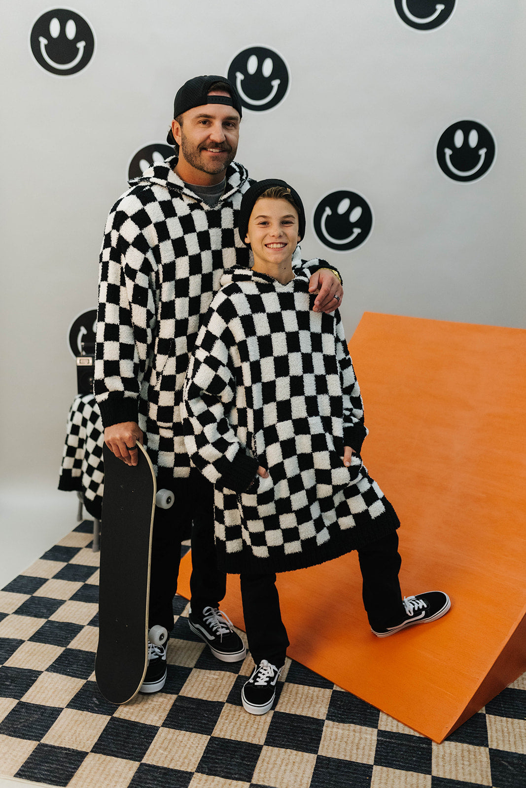 Two people in checkered outfits posing with a skateboard against a wall with smiley face designs.