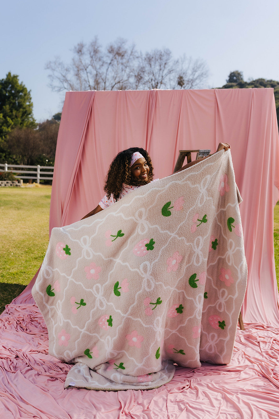 Woman holding a sand blanket with pink and green patterns in front of a pink backdrop outdoors.