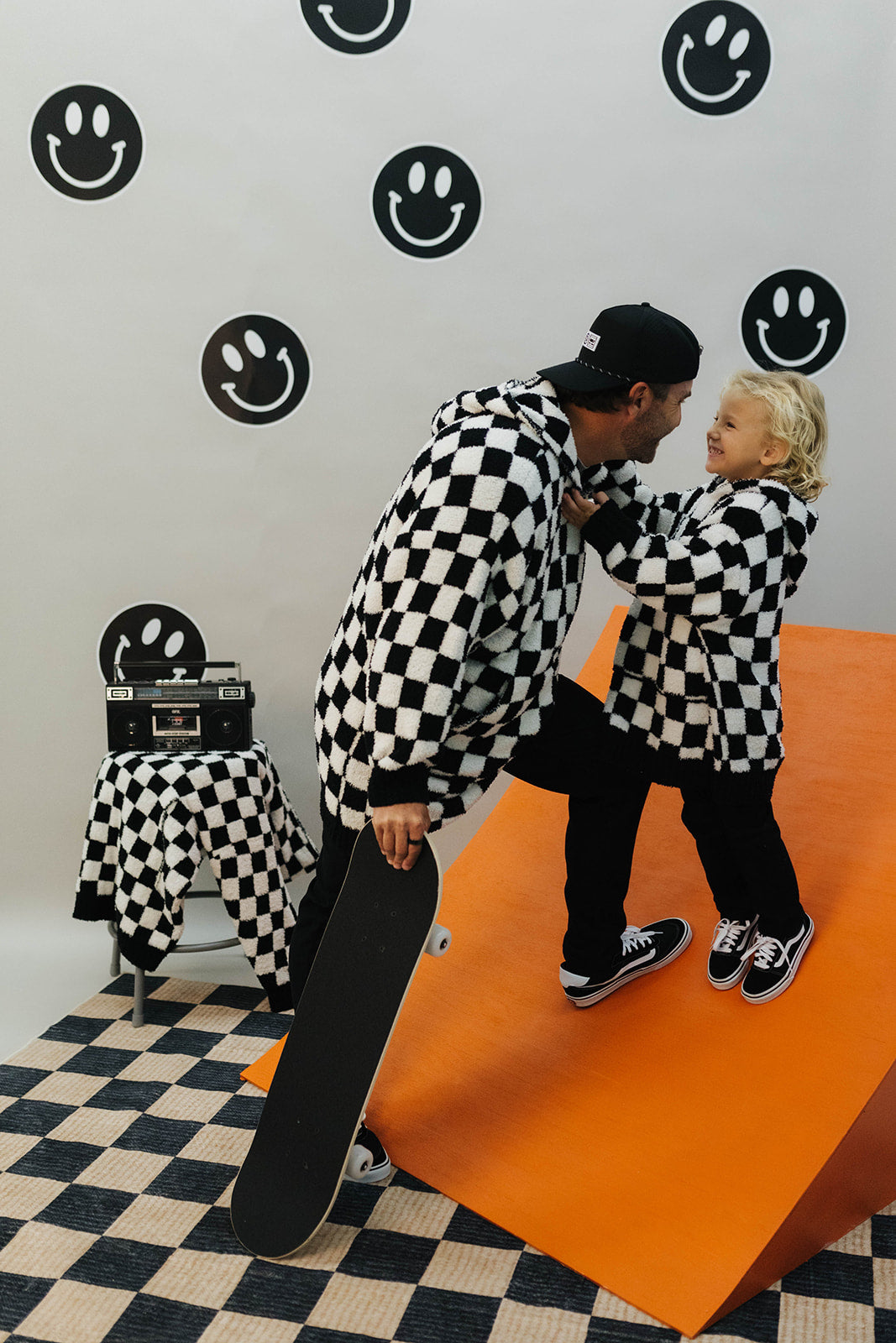 Man and child in checkered outfits with a skateboard on an orange platform against a wall with smiley face patterns.