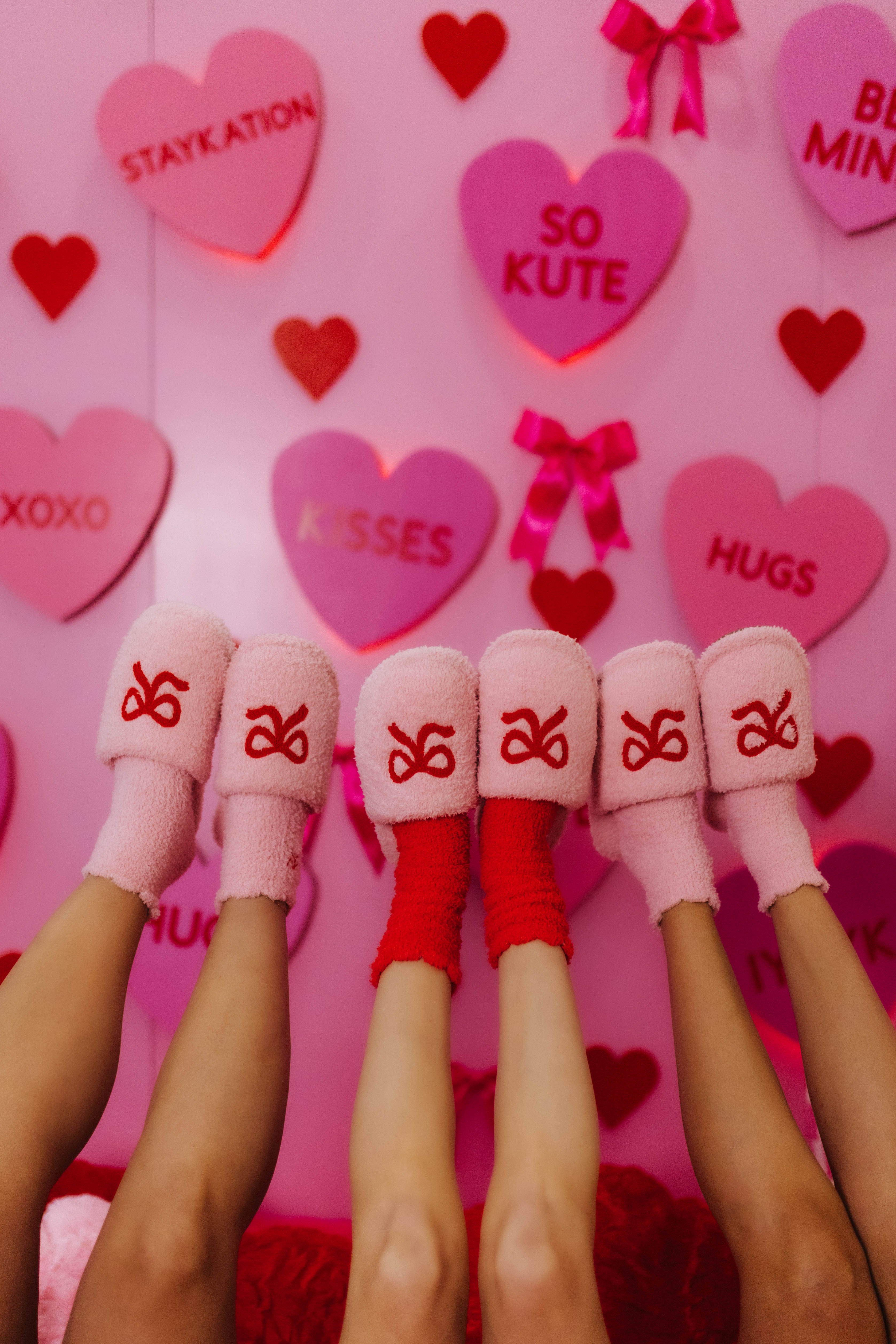 Pink and red socks with slippers in front of a pink background with heart-shaped text and decorations.