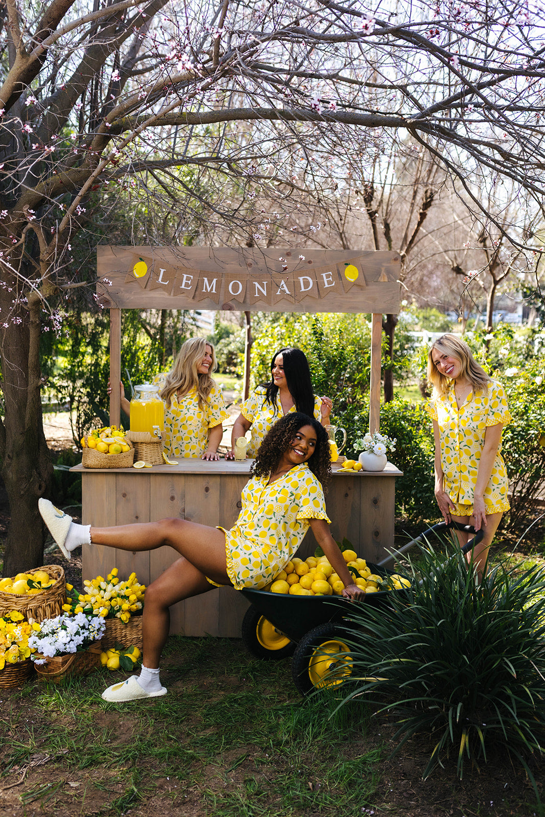 Four women in yellow pjs standing around a lemonade stand with lemonade and lemons in an outdoor setting.
