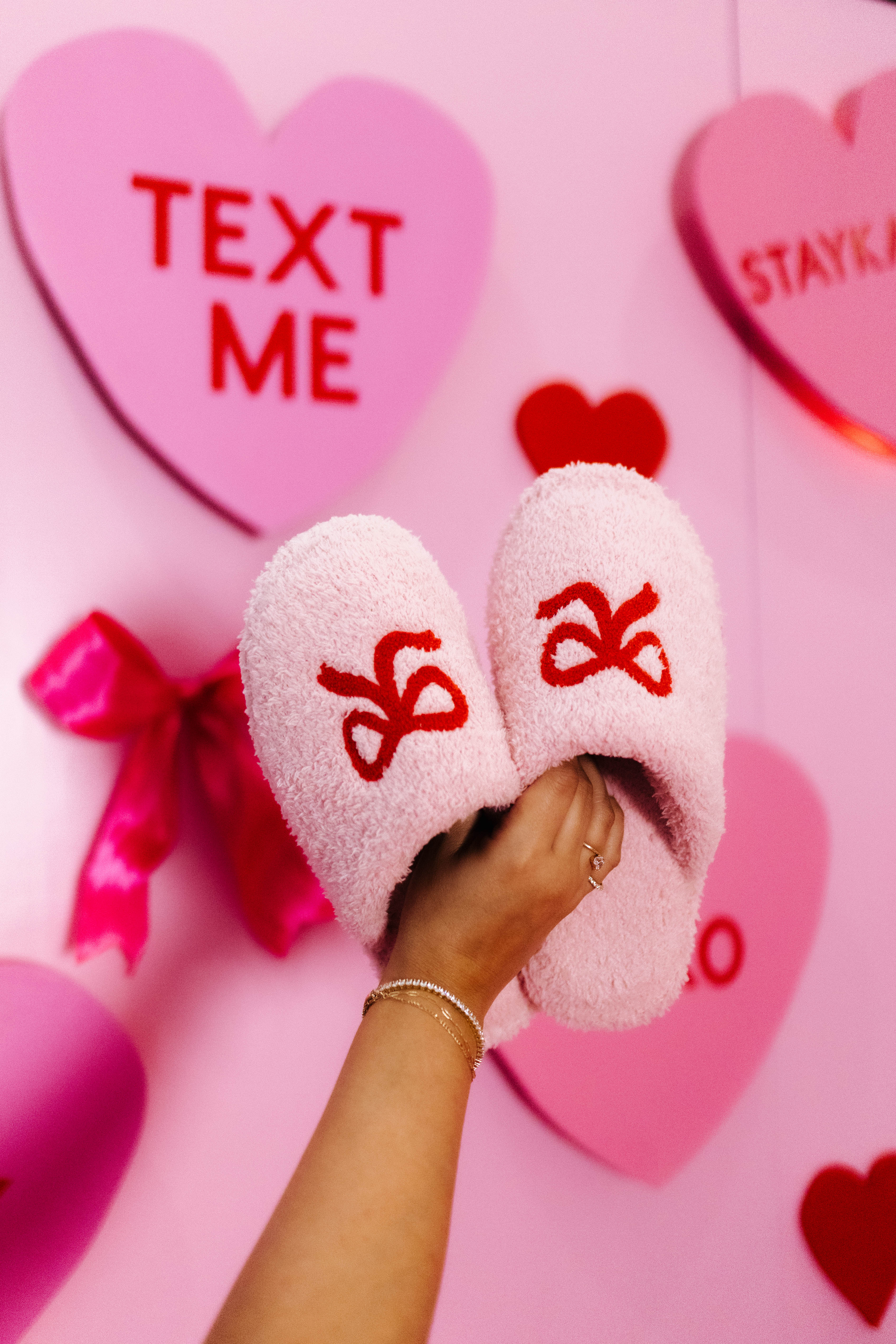 Pink fuzzy slippers with red bow design on a pink background with hearts and text.