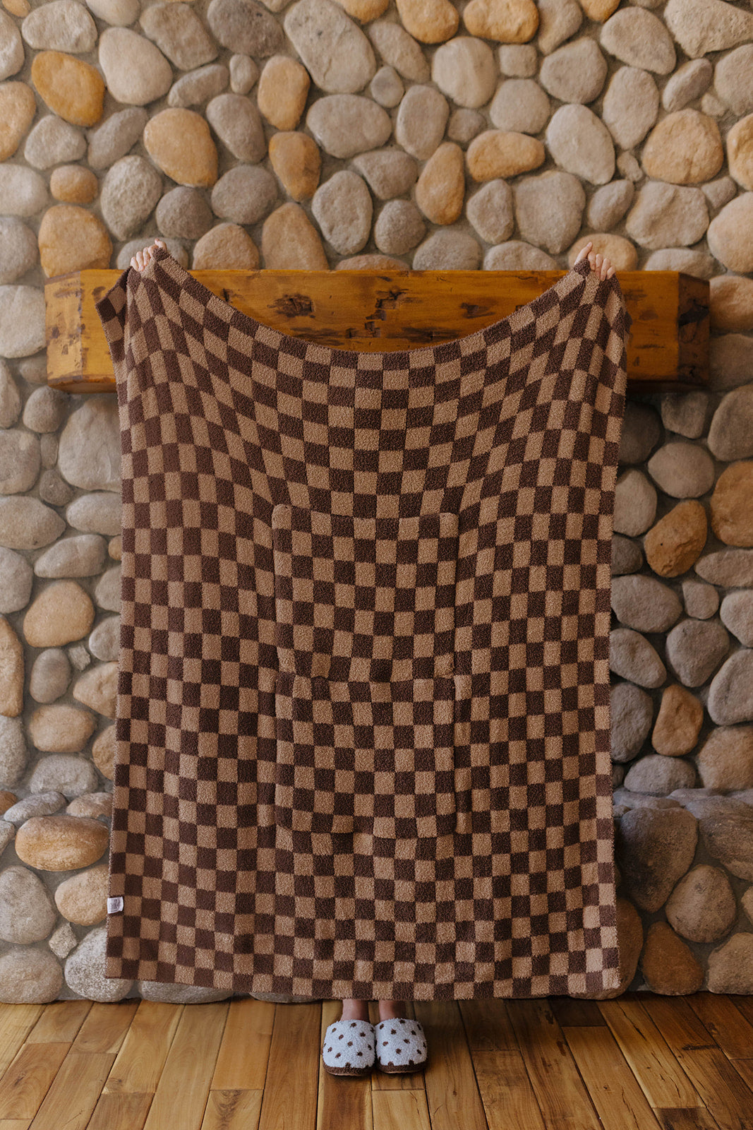 Brown check quillow opened into a blanket in front of a stone fireplace.
