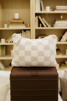 Checkered pillow on a brown suitcase with a bookshelf in the background