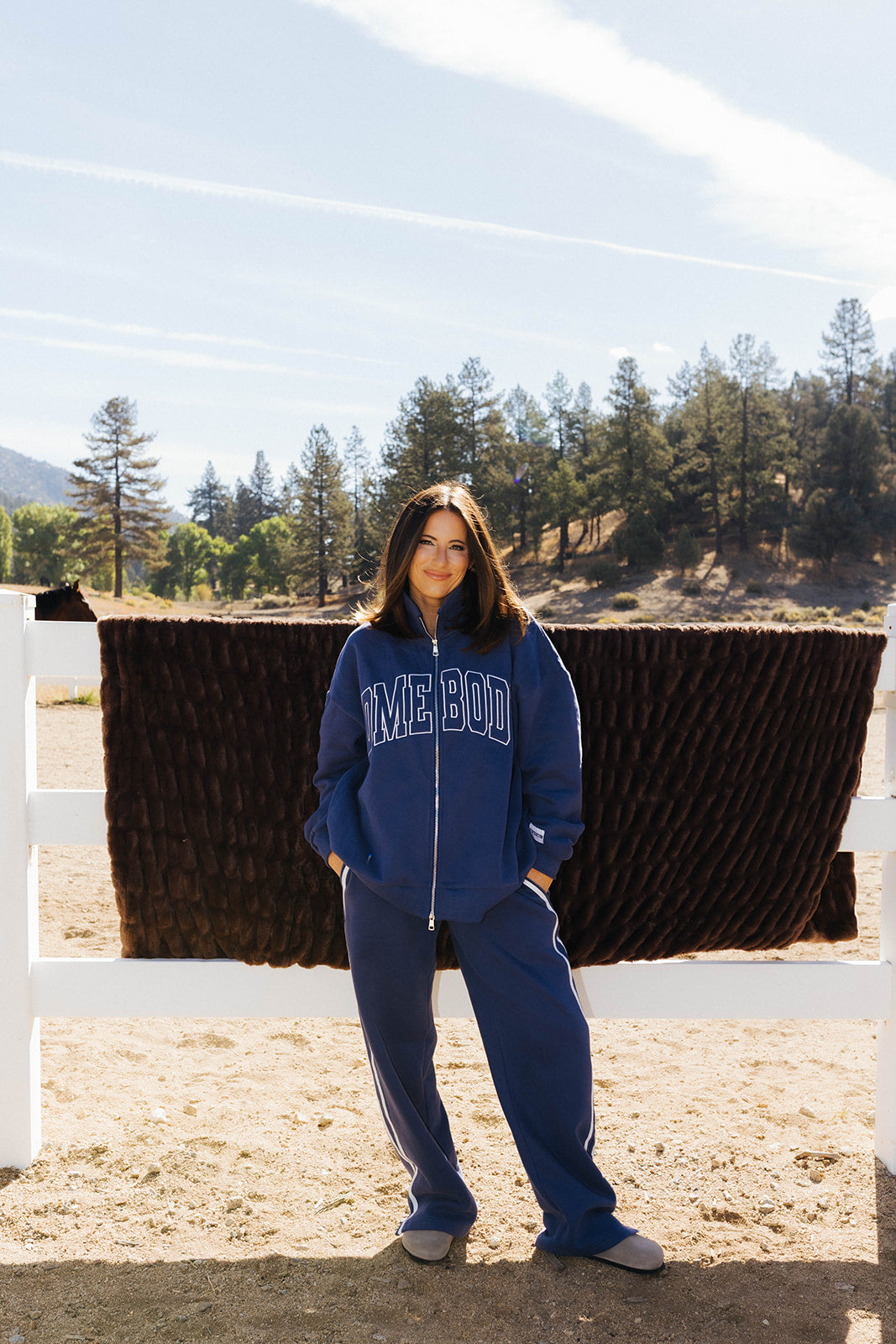 Full length front view of the blue Homebody zip sweatshirt and side stripe sweatpants beside a fence.