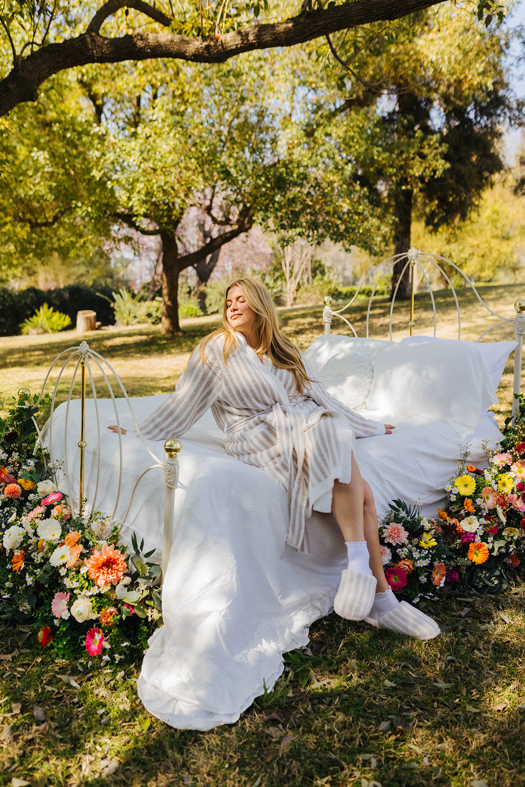 Woman in a white dress sitting on a decorative chair surrounded by flowers and trees.