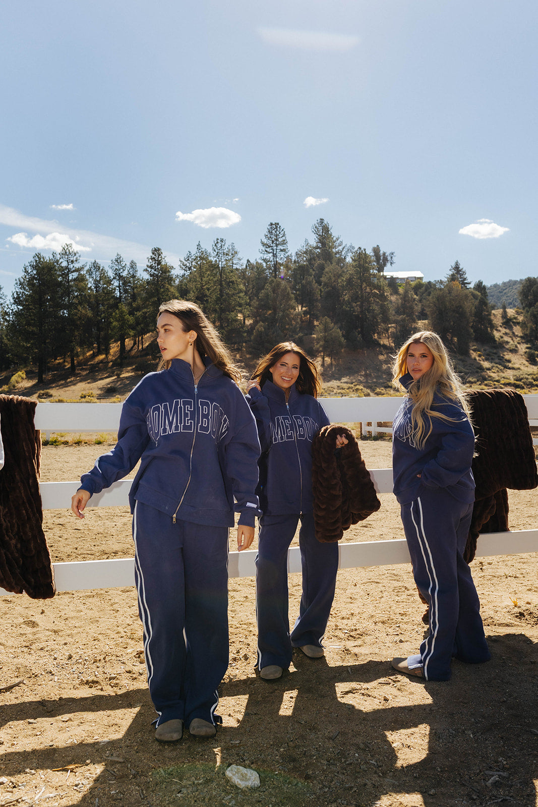 Three models wearing navy Homebody sets beside chocolate brown faux fur blankets draped over a white fence.