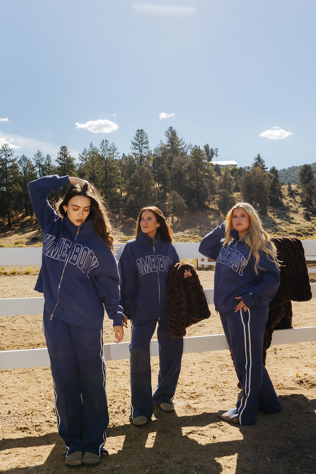 Three models in blue Homebody sweatshirts and sweatpants beside a fence with chocolate brown faux fur blankets.