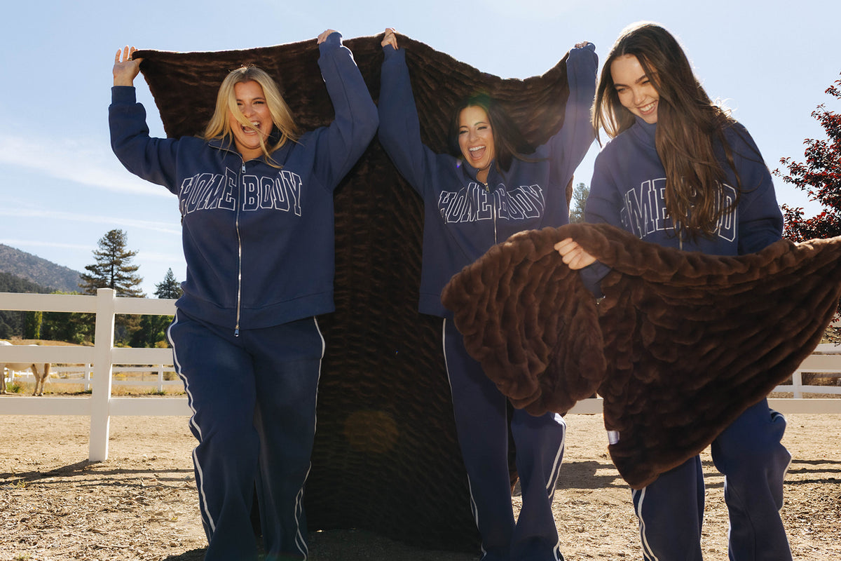 Models holding chocolate brown faux fur blankets open beside a white fence.