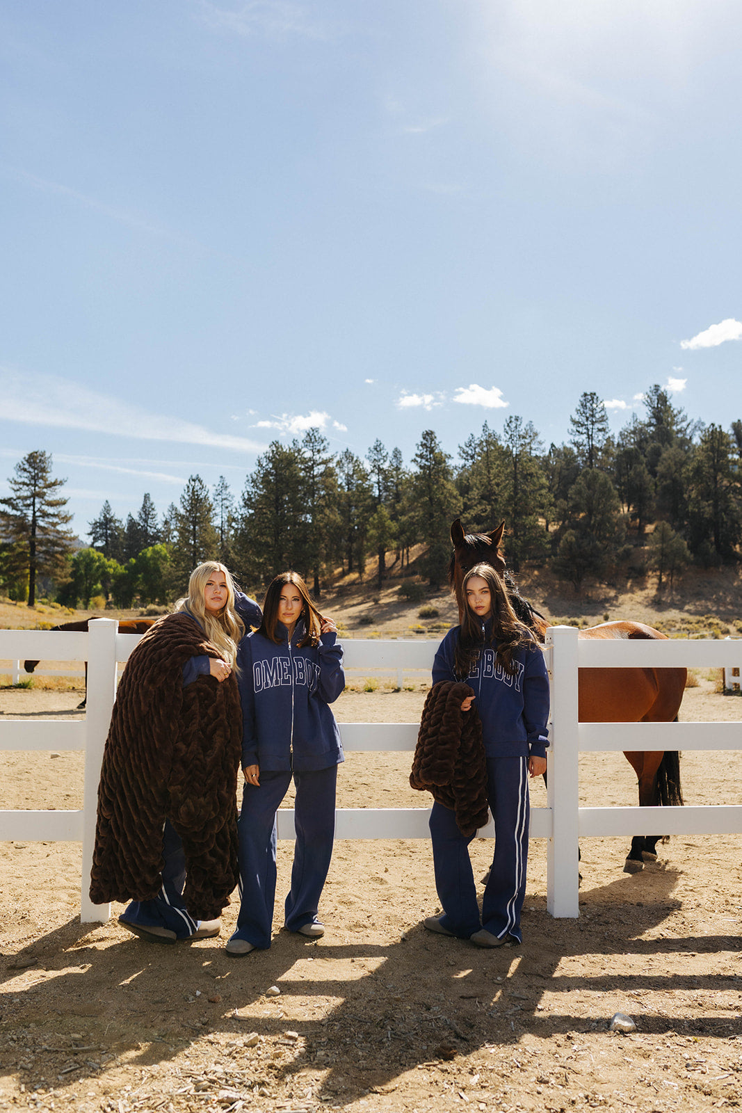 Three models displaying chocolate brown faux fur blankets beside a white fence.