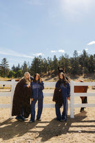 Three models displaying chocolate brown faux fur blankets beside a white fence.